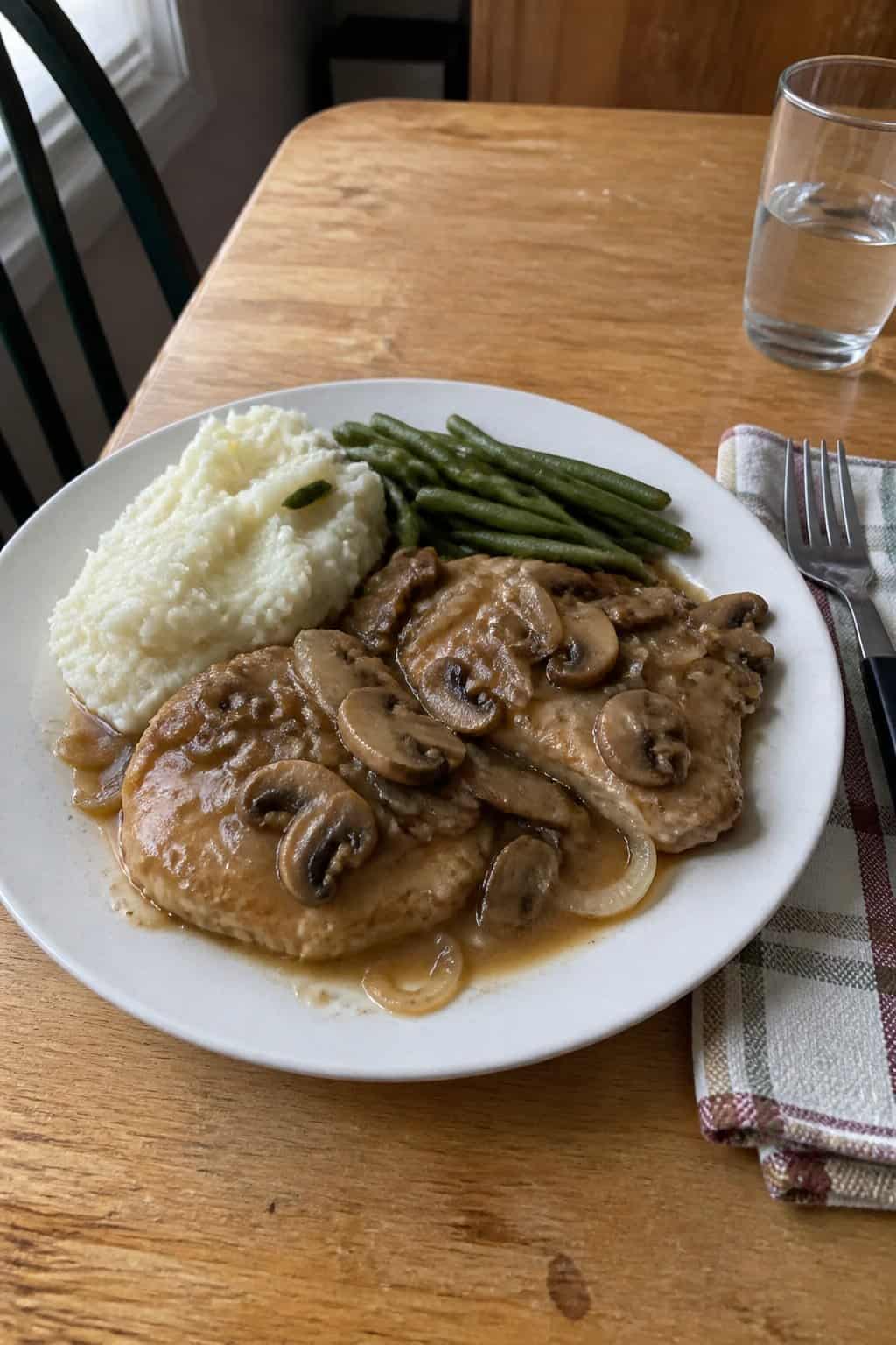A plate of boneless pork chops covered in mushroom and onion sauce with mashed potatoes and green vegetables.