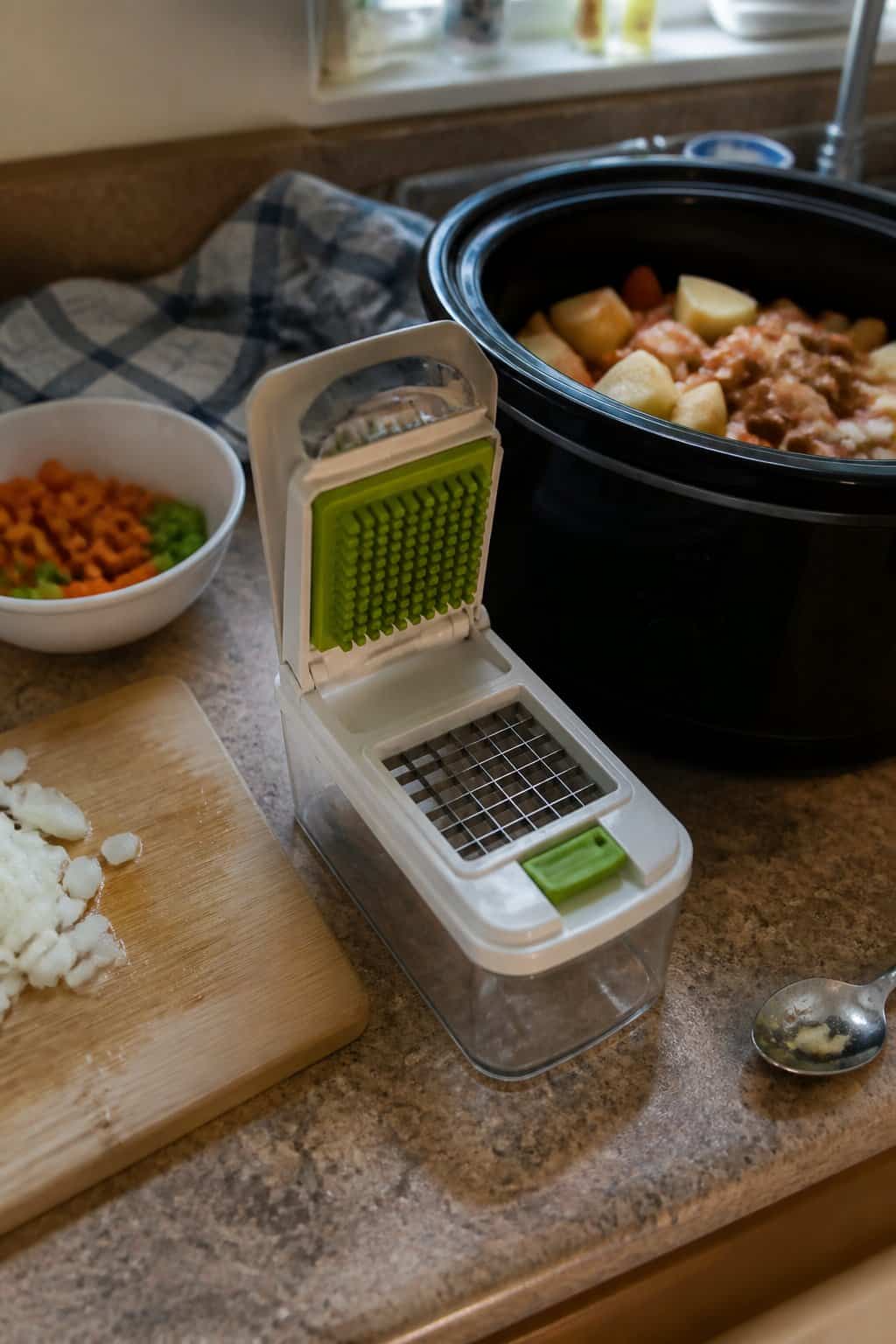 A vegetable chopper sits beside an open slow cooker and bowls of chopped vegetables on a kitchen counter.