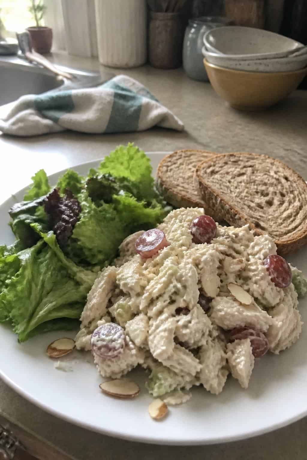 20 Copycat Chicken Recipes for Restaurant Cravings at Home 17 A plate of creamy chicken salad with grapes, almonds, celery, greens, and whole-grain bread on a kitchen counter.