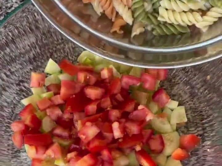 An overhead view shows cooked rotini pasta in a bowl next to a bowl of chopped cucumbers and tomatoes.