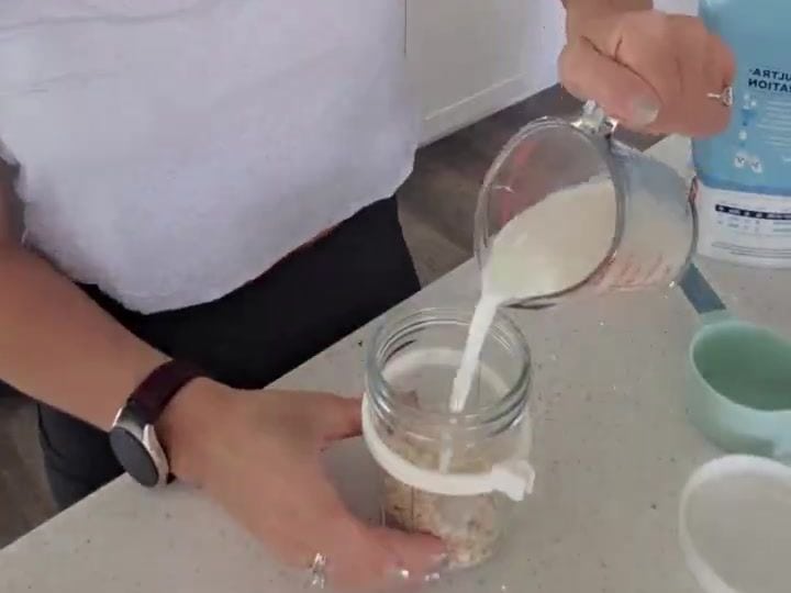 Creamy High-Protein Overnight Oats With Fairlife Milk 4 Fairlife milk is being poured in a stream into a glass jar containing oats with a spoon resting inside.