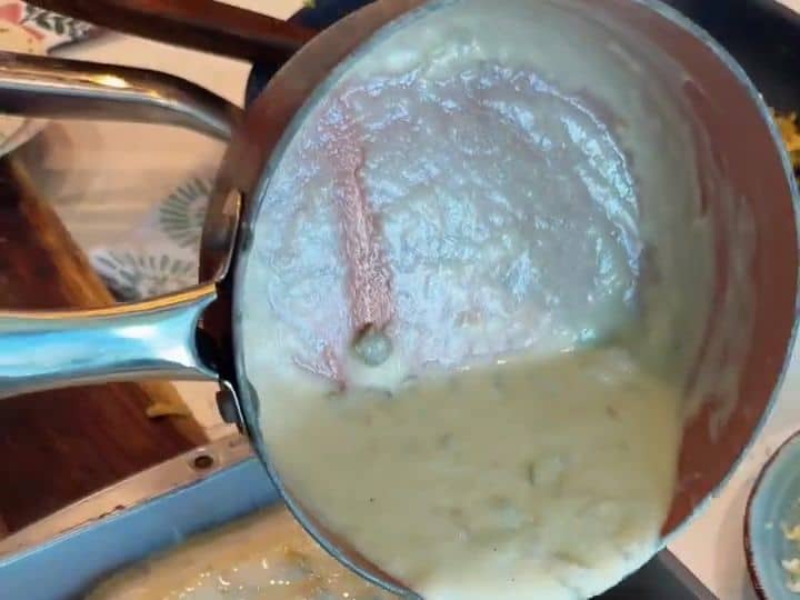 Creamy white green chile sauce is being poured from a pot over rolled tortillas lined up in a baking dish.
