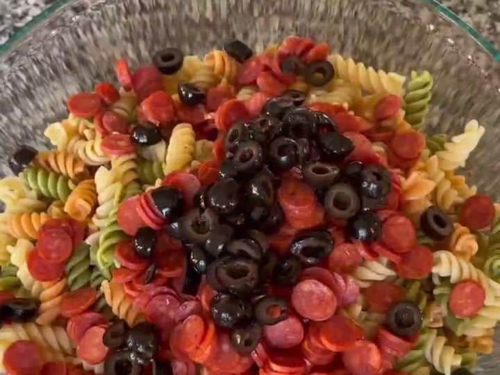 Sliced black olives are being poured over a bowl of pasta salad mixed with pepperoni.