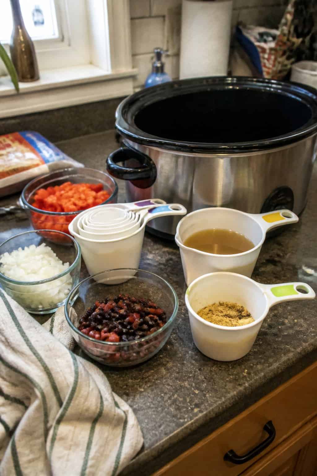A measuring cup set and prepared ingredients sit beside a slow cooker on a kitchen counter.