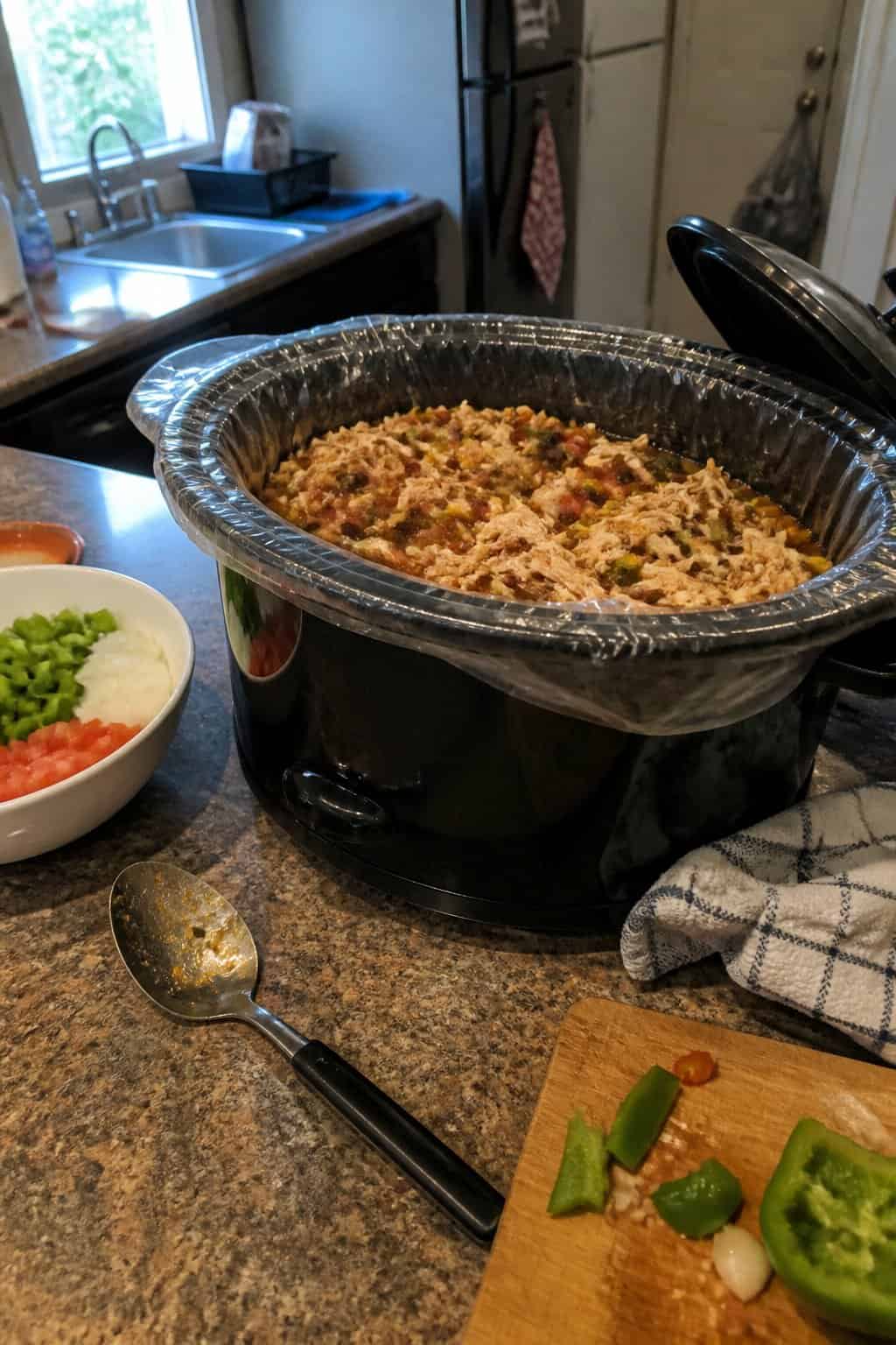 An open slow cooker with a disposable liner, a homemade meal, and prepared ingredients on a kitchen counter.