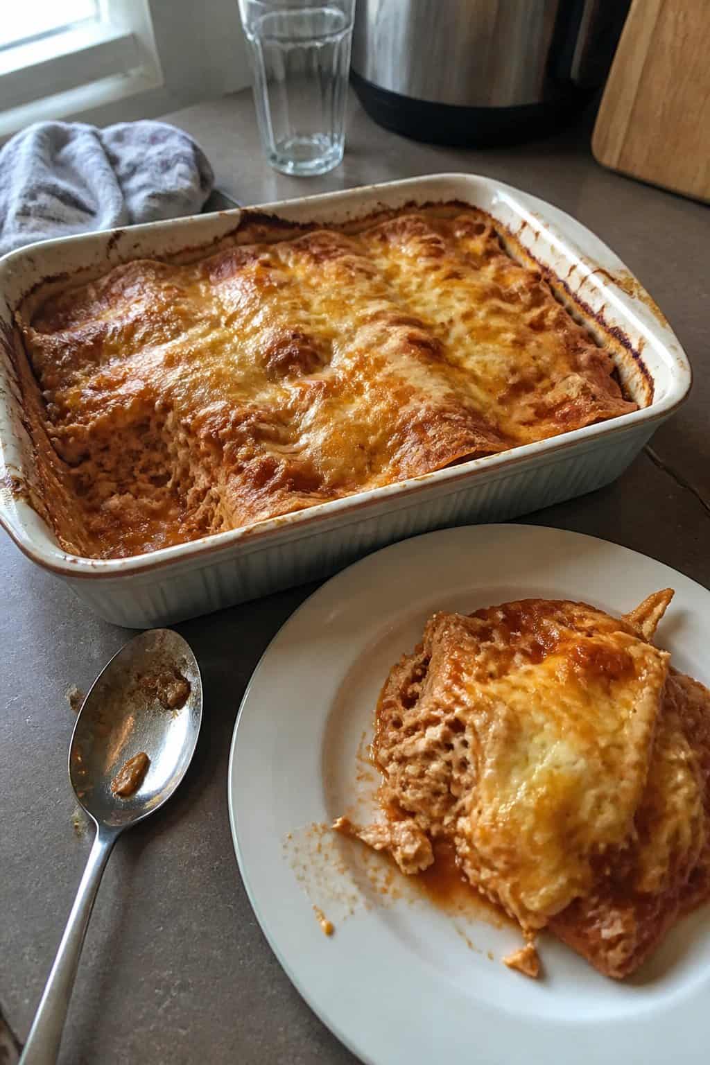 A homemade chicken enchilada casserole in a baking dish beside a serving spoon and a plated portion.