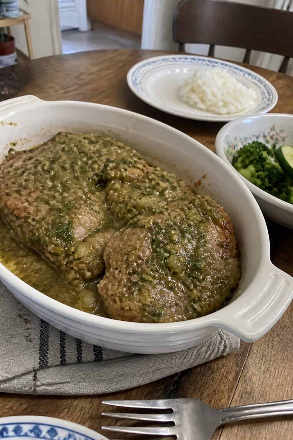 Tender pork chops covered in green salsa verde are served with rice and vegetables on a kitchen table.