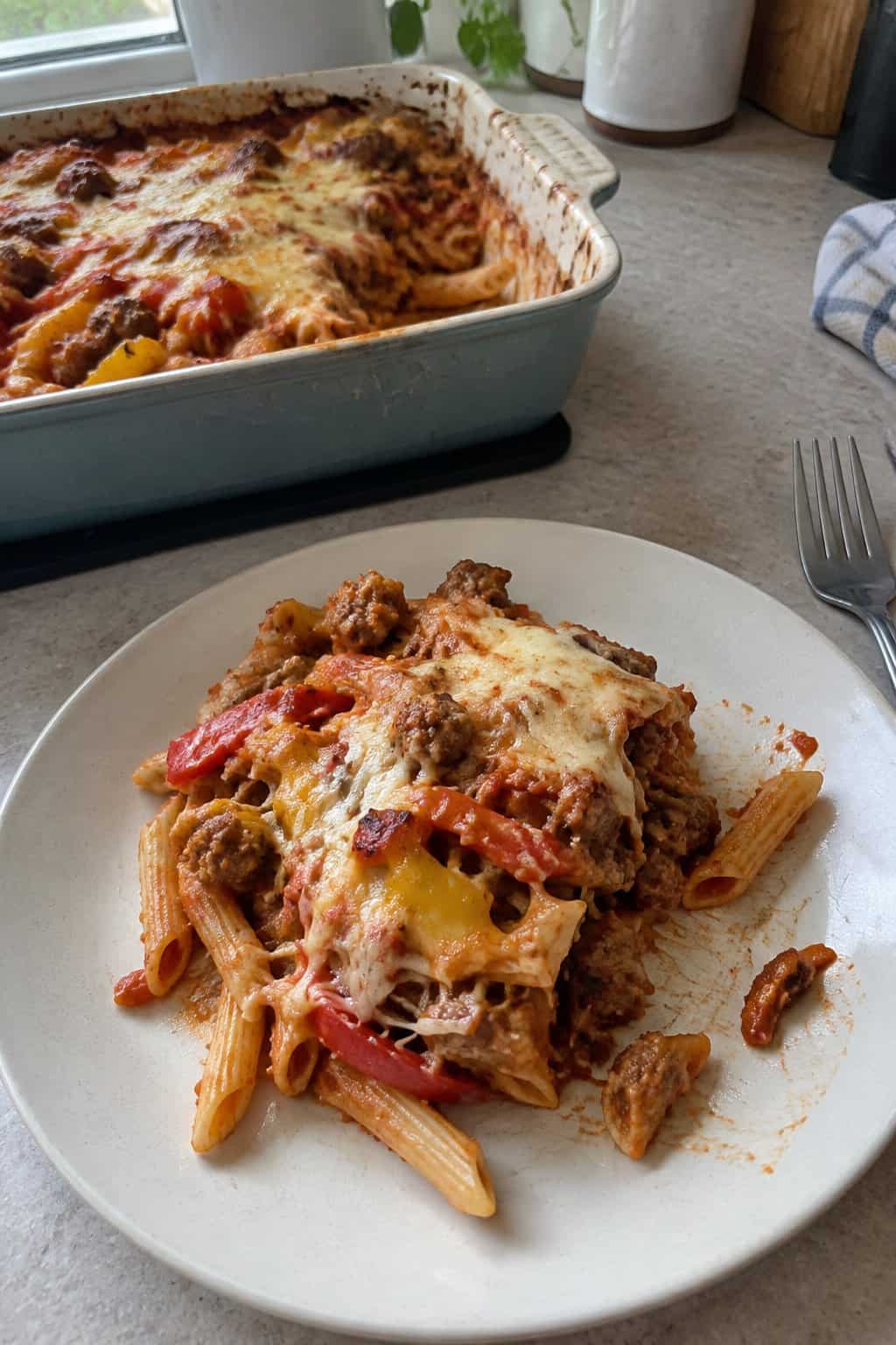 A serving of sausage and pepper penne bake with melted cheese on a plate beside the baking dish.