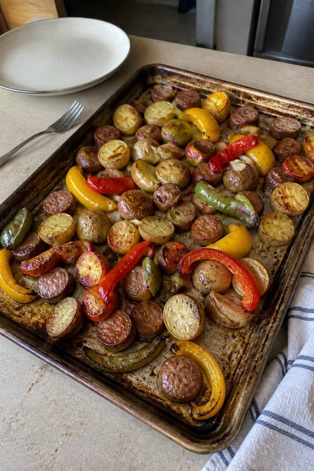 A sheet pan filled with roasted sausage, potatoes, and colorful bell peppers on a kitchen counter.