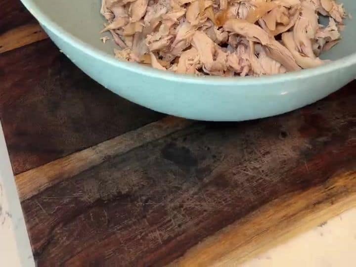 A bowl filled with shredded rotisserie chicken meat sitting on a wooden cutting board.