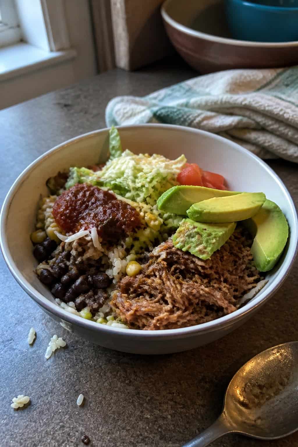 A bowl of shredded beef taco ingredients served over rice with beans, corn, tomatoes, lettuce, cheese, avocado, and salsa on a kitchen table.