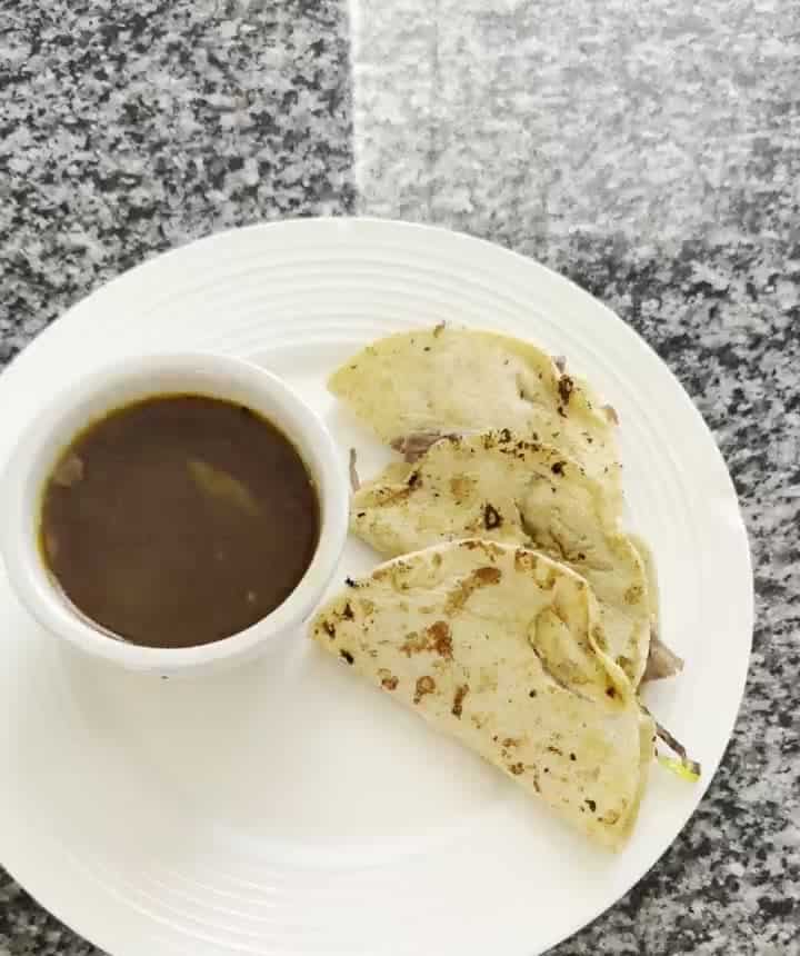 Three crispy birria street tacos on a white plate with a cup of consomme broth for dipping