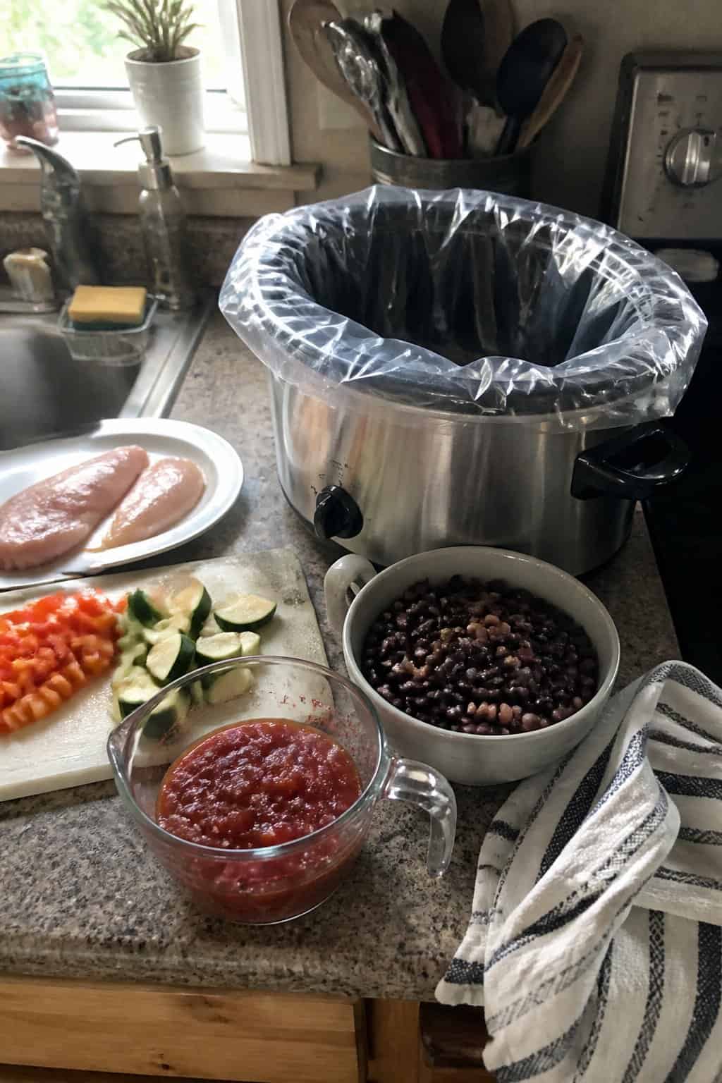 A slow cooker with a disposable liner being prepared on a kitchen counter beside ingredients and cooking utensils.