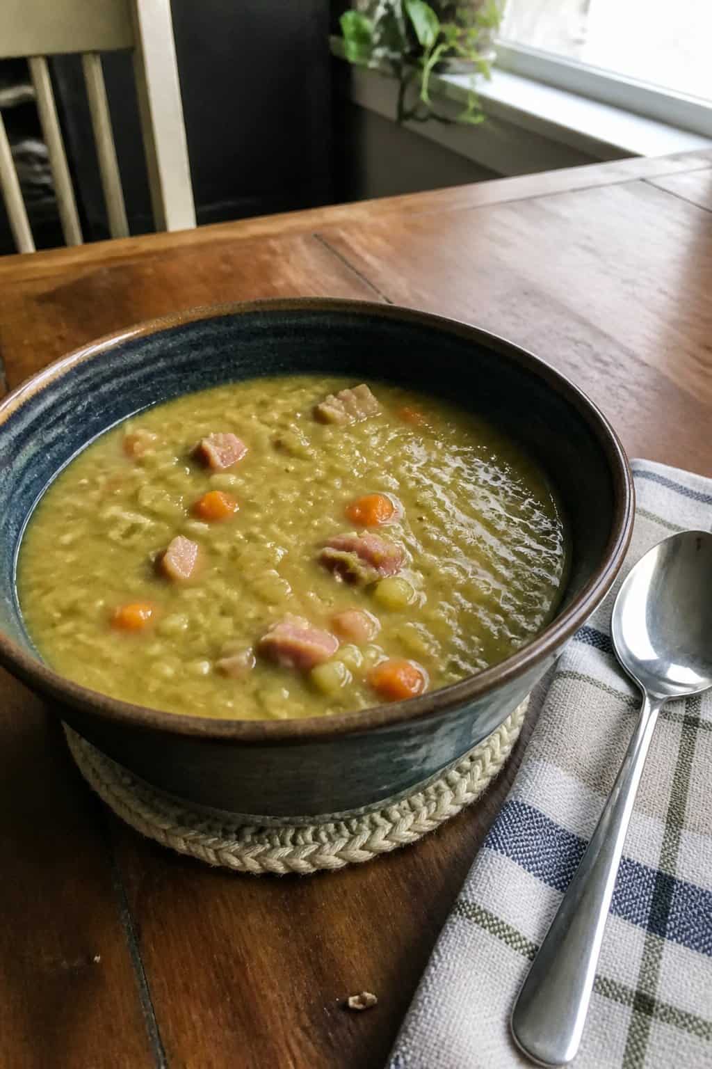 Ham Soup Recipes for Chilly Nights 6 A bowl of thick split pea soup with diced vegetables and pieces of ham on a kitchen table beside a spoon.
