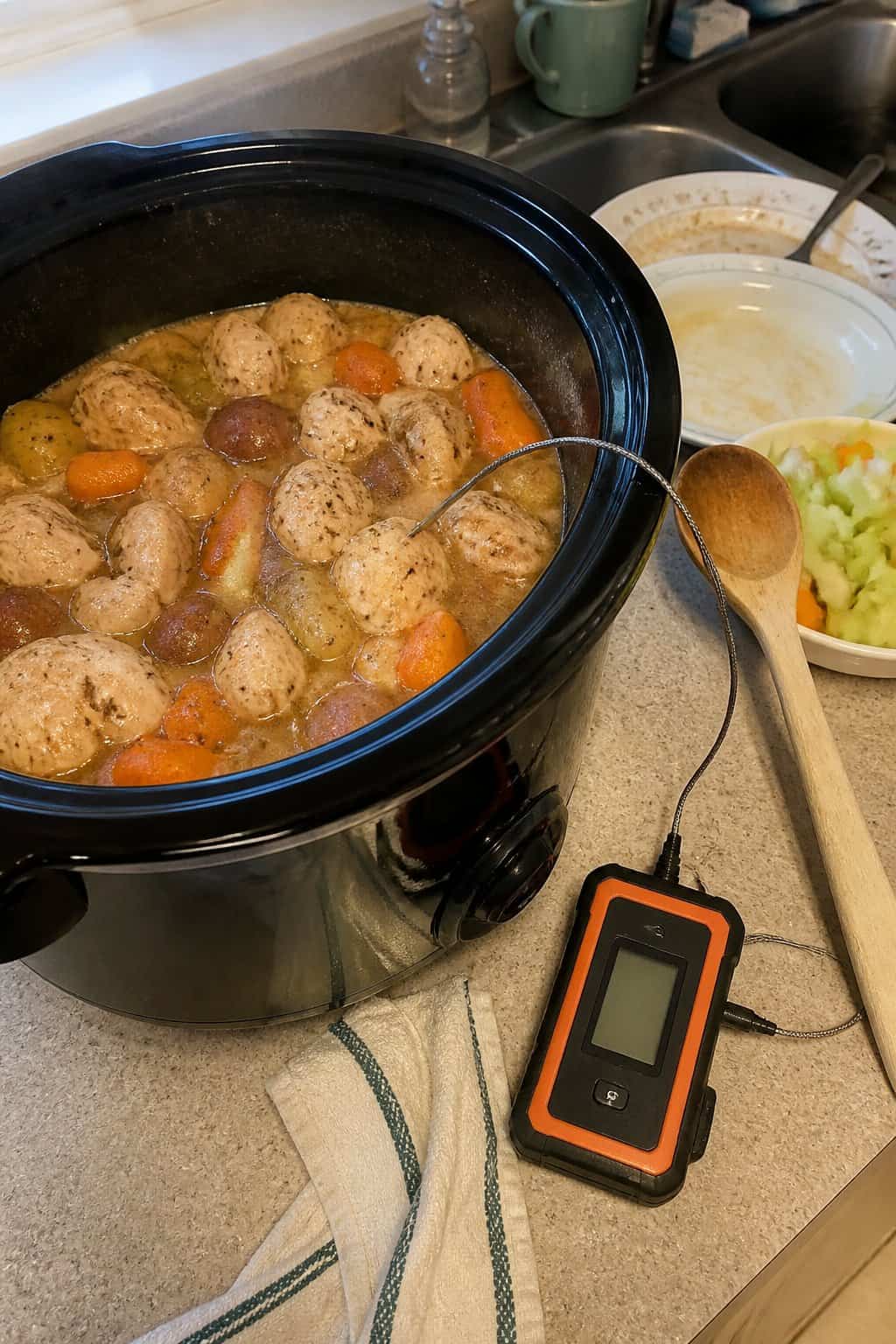 A digital meat thermometer checks a meal in an open slow cooker on a kitchen counter.