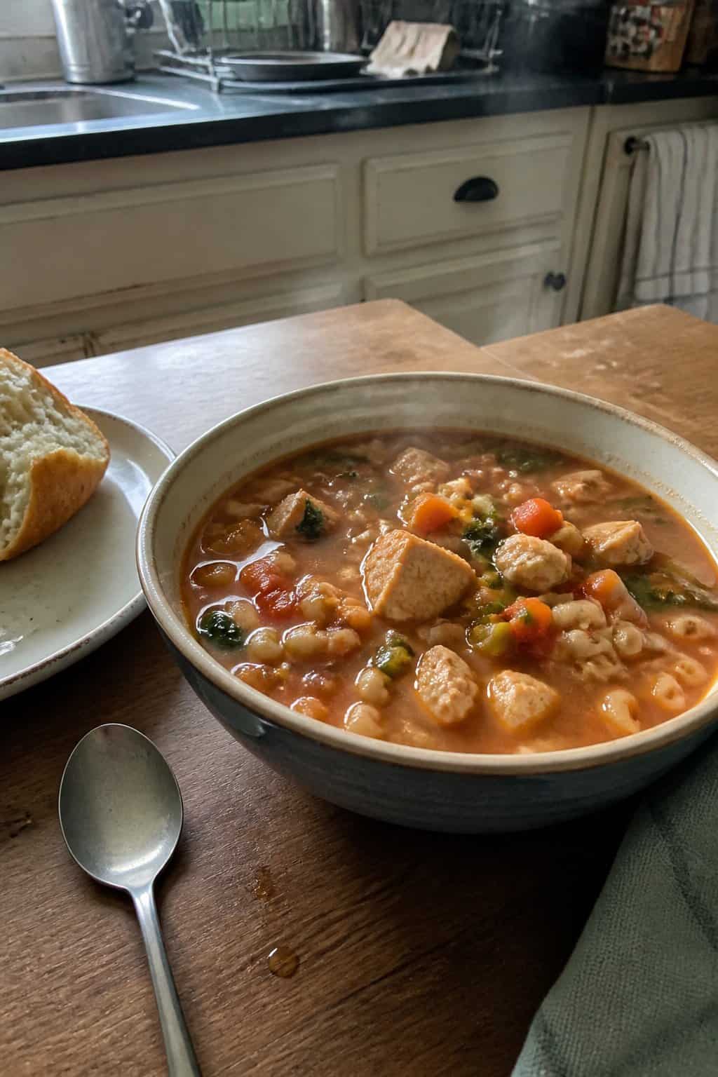 25 Turkey Soup Recipes That Make the Whole House Smell Amazing 18 A bowl of turkey minestrone soup with pasta, vegetables, and bread on a kitchen table.
