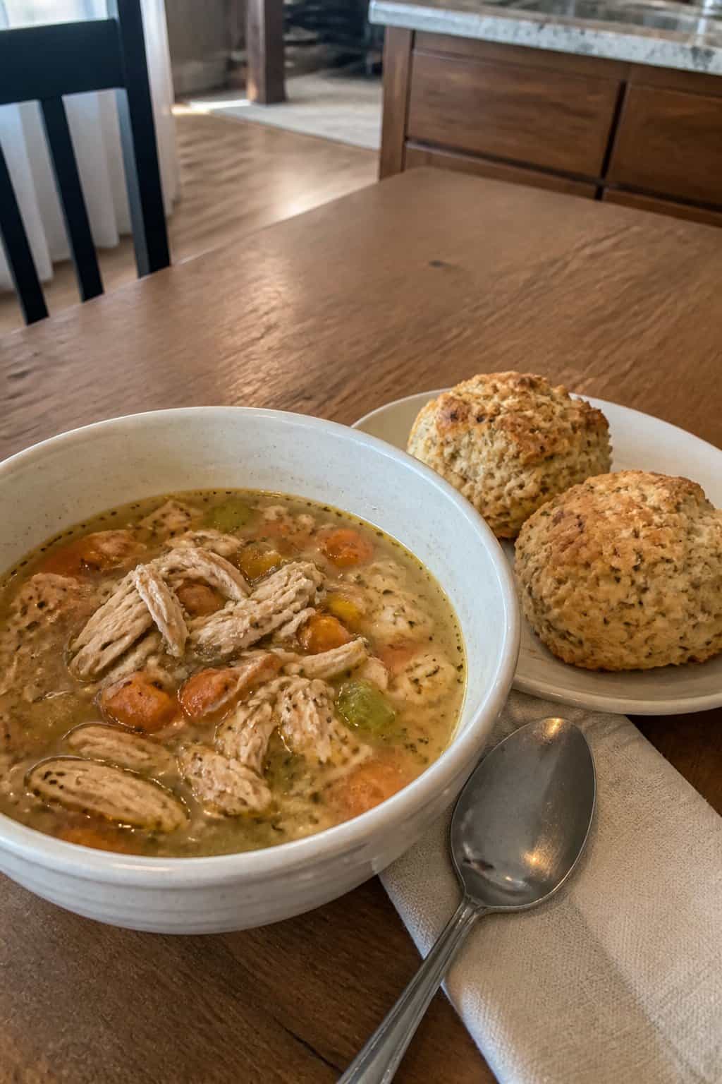 25 Turkey Soup Recipes That Make the Whole House Smell Amazing 24 A bowl of turkey soup with vegetables sits beside golden garlic herb biscuits on a kitchen table.