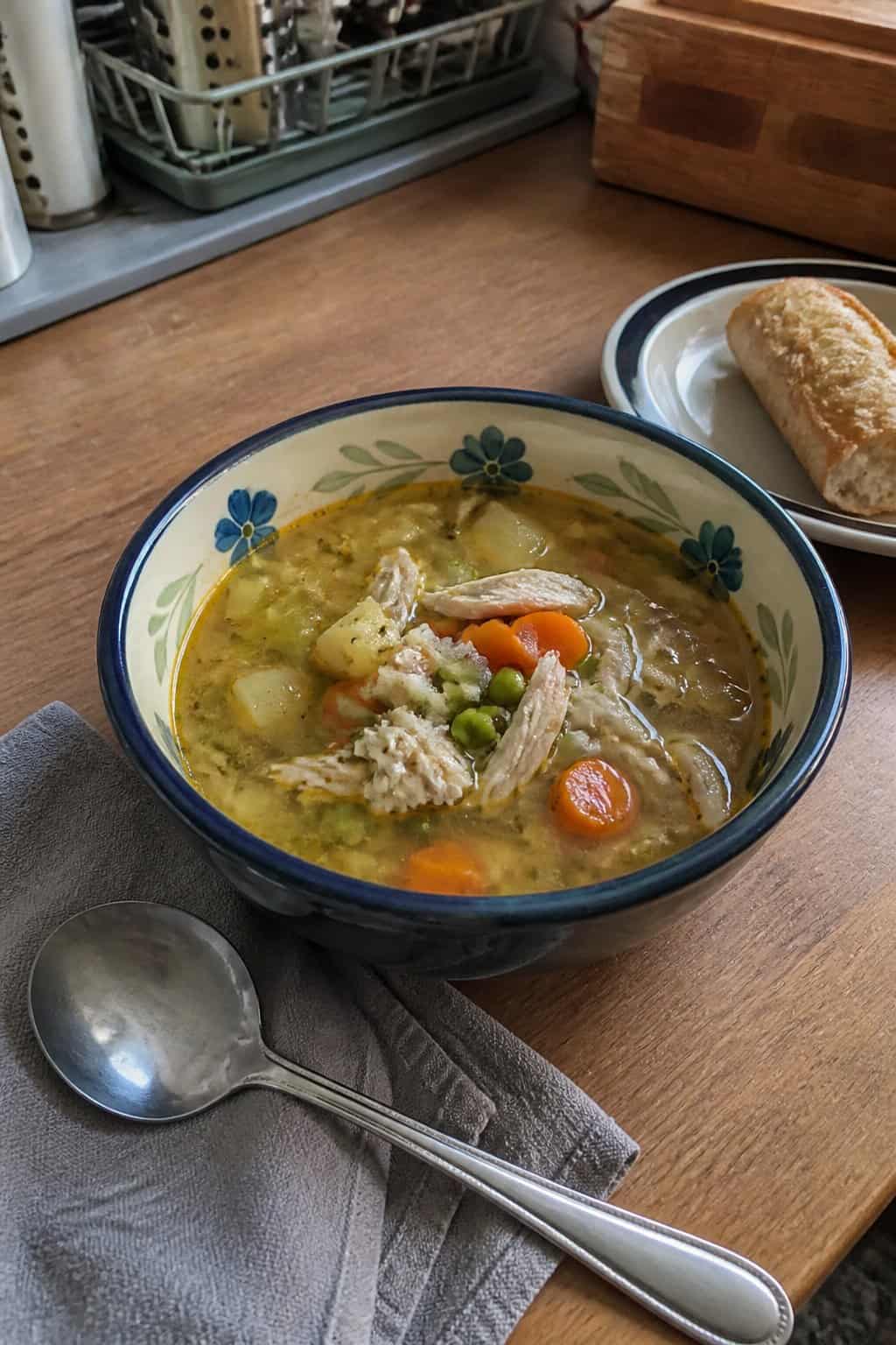 25 Turkey Soup Recipes That Make the Whole House Smell Amazing 23 A bowl of homemade turkey vegetable soup with carrots, celery, peas, and potatoes on a kitchen table beside bread and a spoon.