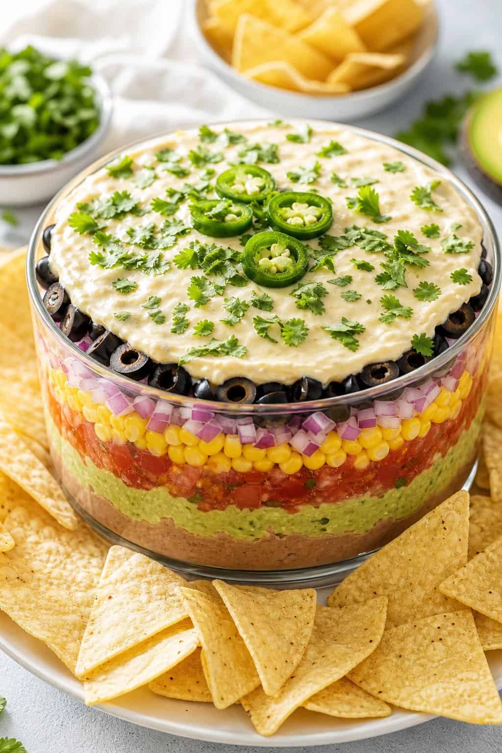 A colorful seven-layer vegan dip in a glass dish surrounded by tortilla chips.