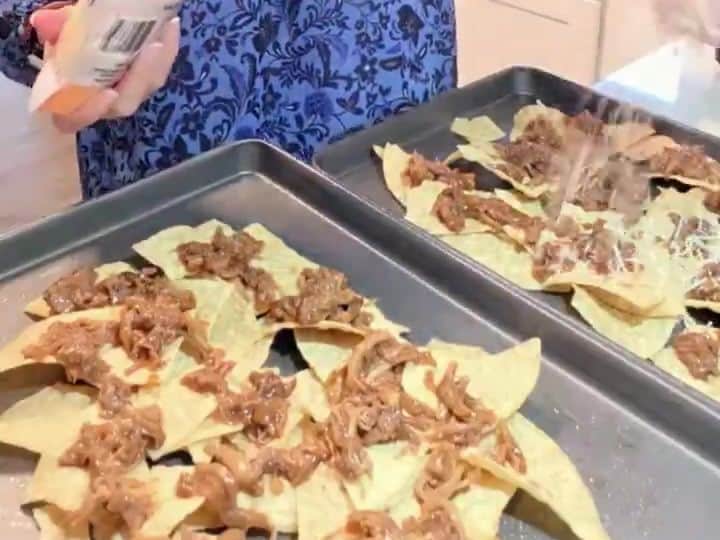 A tray of tortilla chips topped with shredded pulled pork is being sprinkled with shredded cheese from a bag.