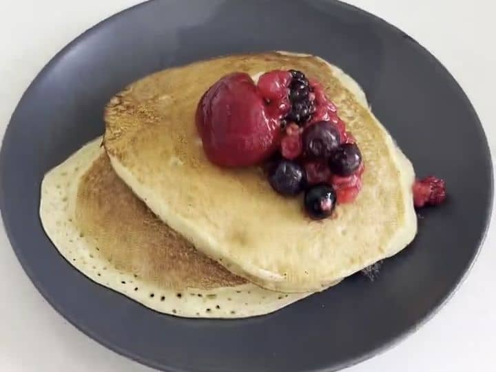 A stack of golden pancakes topped with a mix of thawed berries sits on a dark plate.