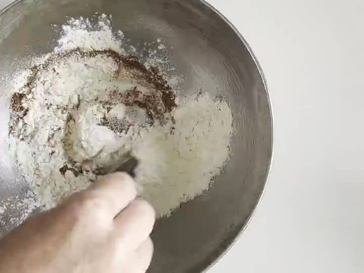 A hand stirs a metal bowl where a wet mixture is being combined into dry flour with a spoon.